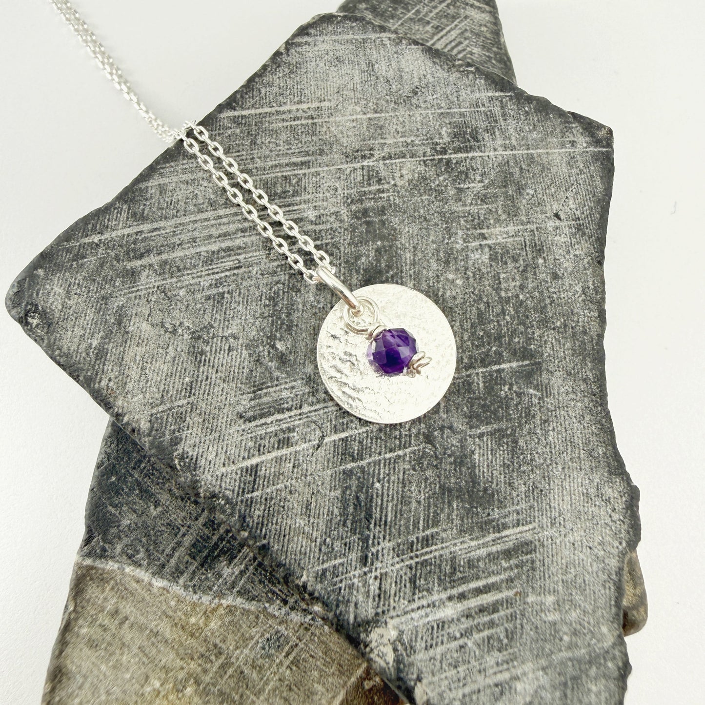 Amethyst Birthstone Charm Pendant Necklace in Sterling Silver on a piece of gray stone.