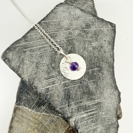 Amethyst Birthstone Charm Pendant Necklace in Sterling Silver on a piece of gray stone.