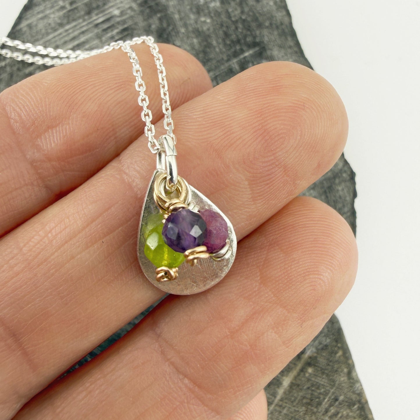 Silver necklace with a teardrop-shaped pendant featuring purple and green stones, held in a hand against a stone background.