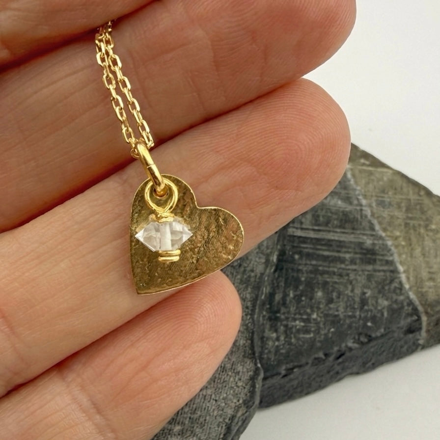 Gold heart-shaped necklace with a diamond birthstone charm held by a hand against a stone background