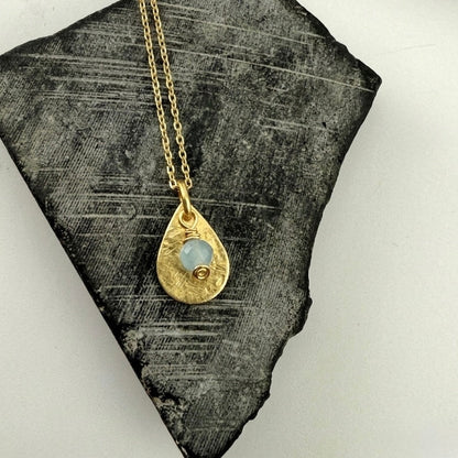 Gold necklace with a teardrop-shaped pendant and aquamarine charm on a textured stone surface