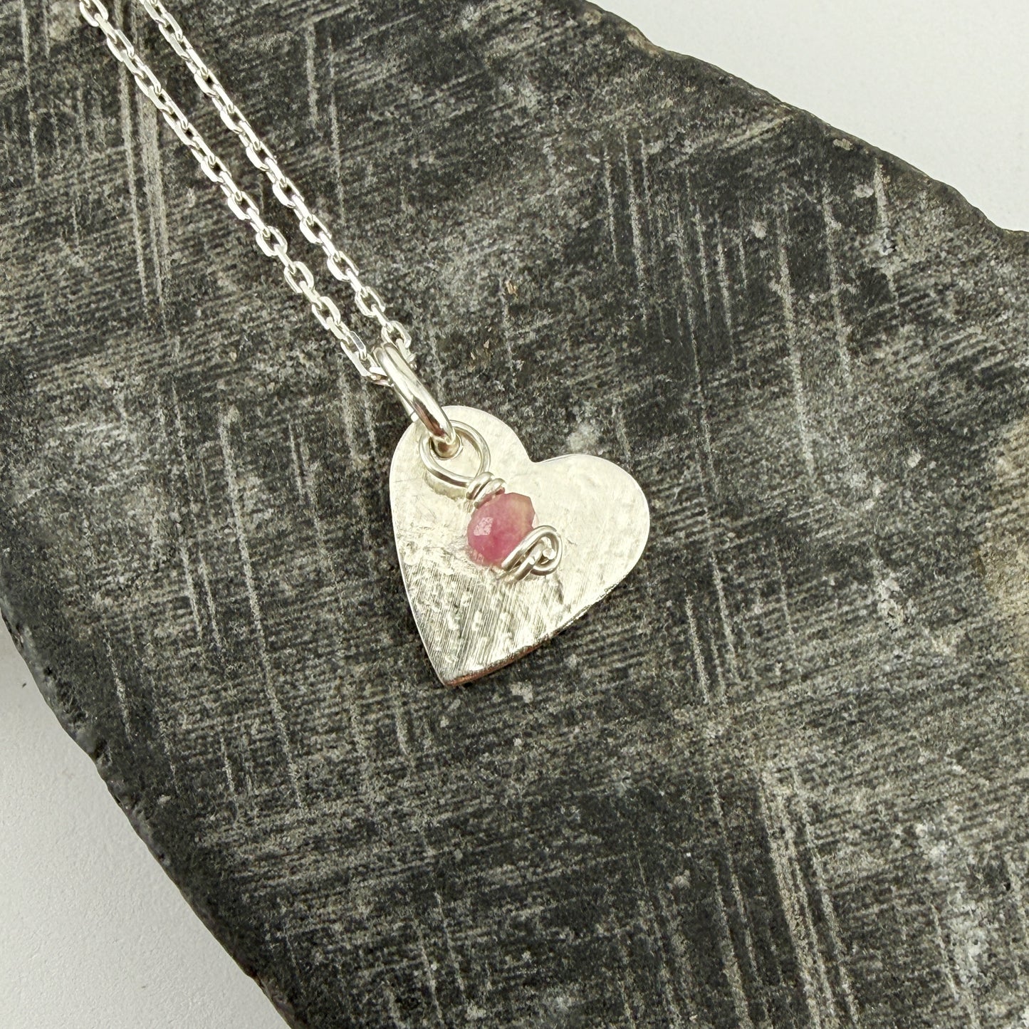 Silver heart-shaped pendant with a tourmaline gemstone charm on a textured stone surface