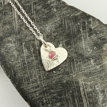 Silver heart-shaped pendant with a tourmaline gemstone charm on a textured stone surface