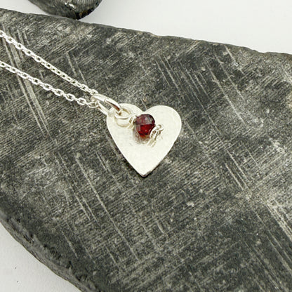 Silver heart-shaped necklace with a January garnet birthstone charm on a textured stone surface